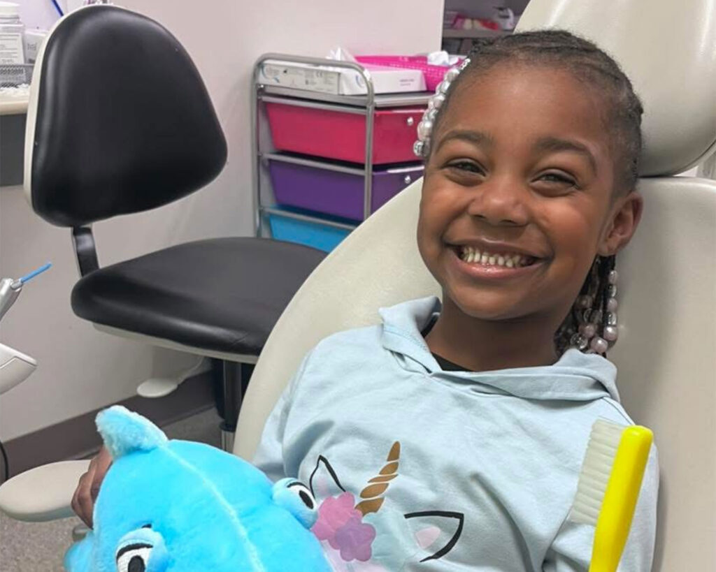 young child in dentist chair holding a stuffed shark with a big smile on her face